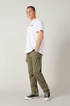 A person stands against a light gray background wearing a white t-shirt, 686 Men's Everywhere® Pant - Relaxed Fit in olive green, and black-and-white sneakers, gazing slightly to the side with one hand in their pocket.
