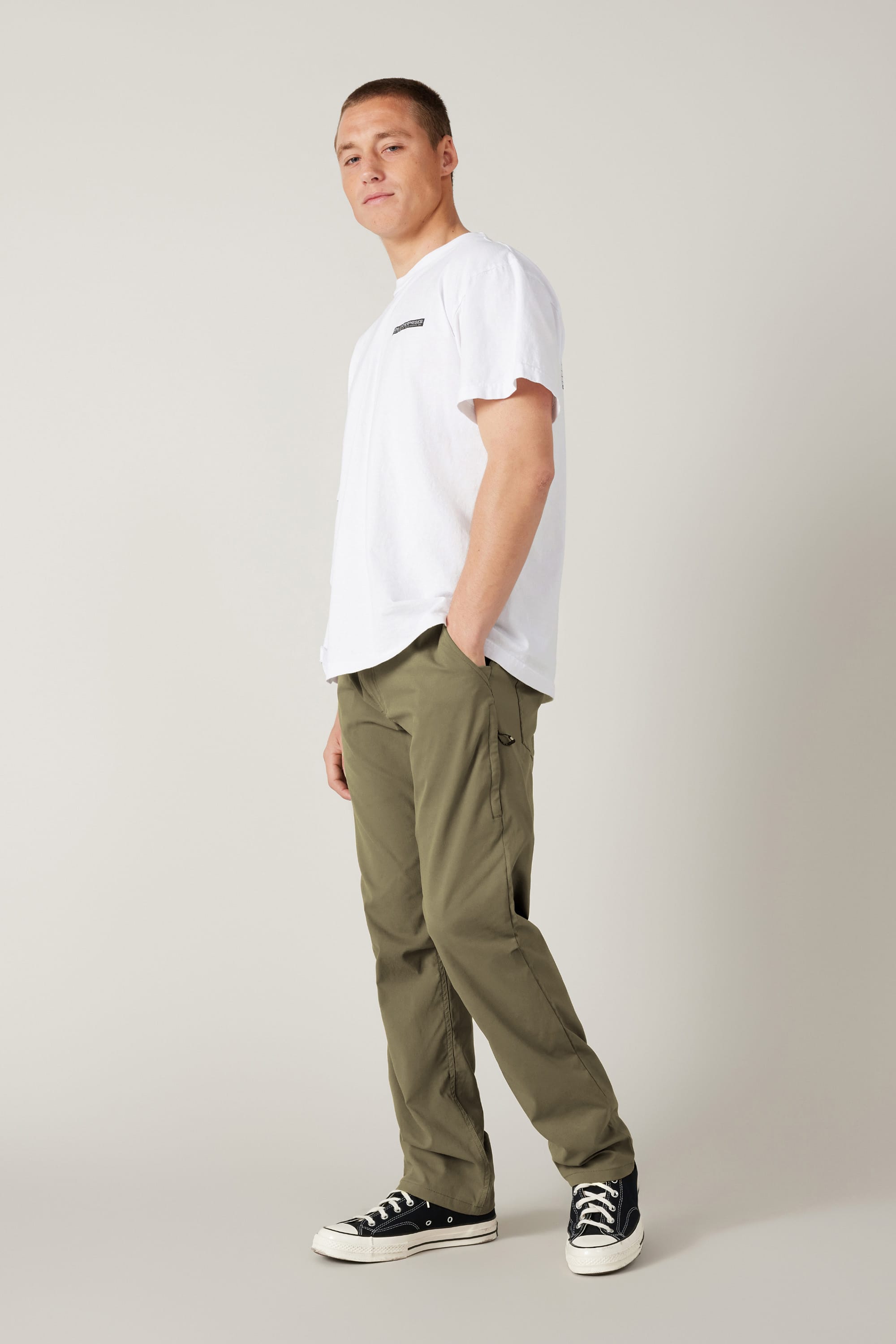 A person stands against a light gray background wearing a white t-shirt, 686 Men's Everywhere® Pant - Relaxed Fit in olive green, and black-and-white sneakers, gazing slightly to the side with one hand in their pocket.