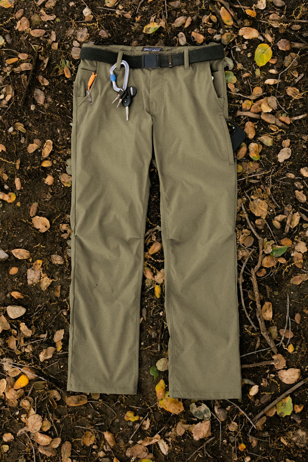 The 686 Men's Everywhere® Pant - Relaxed Fit in olive green features stain-resistant fabric and tool-ready waistband, shown with a black belt and orange pen, laid on forest floor—perfect for outdoor work and easy cleaning.