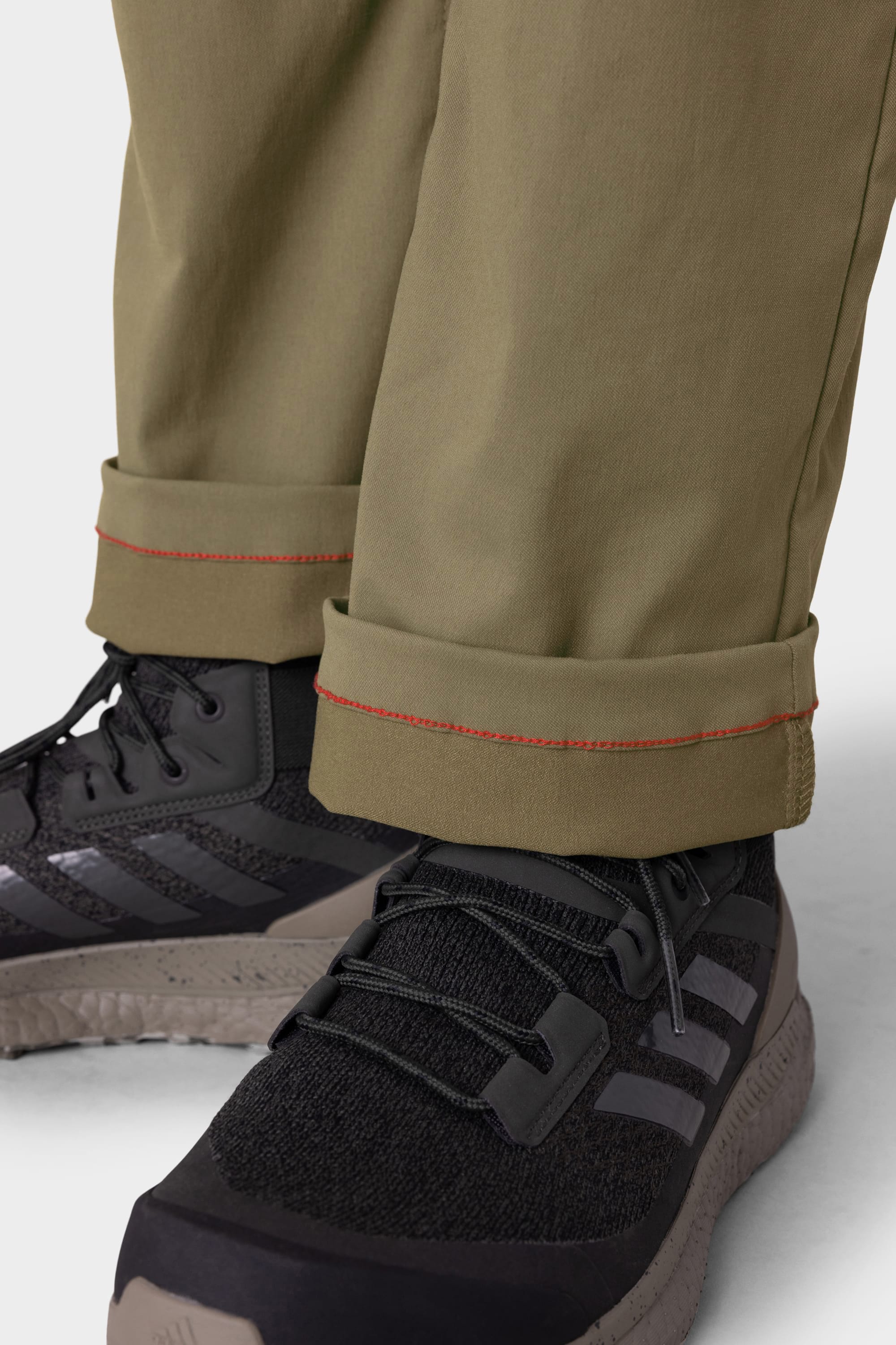 Close-up of a person wearing 686 Men's Everywhere® Pant - Relaxed Fit in olive green with red stitching, paired with black athletic shoes featuring light gray soles and three gray stripes. Brand: 686.