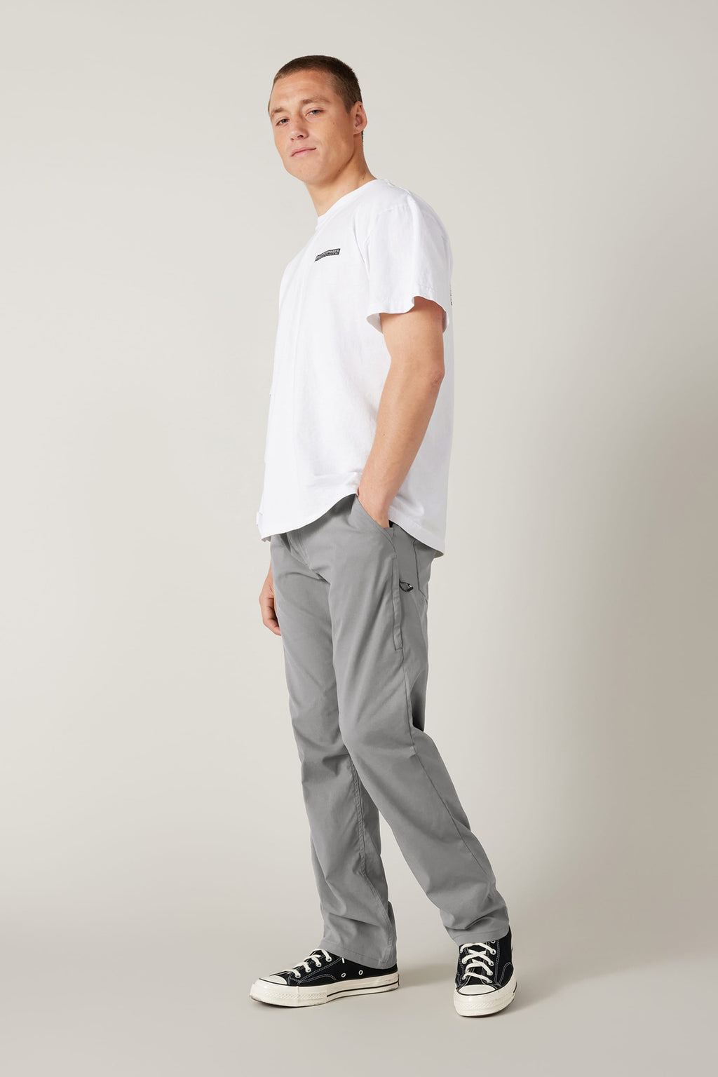 A person stands against a light gray background, wearing a white t-shirt, black-and-white sneakers, and 686 Men's Everywhere® Pant - Relaxed Fit from 686, with one hand in their pocket and a relaxed expression.