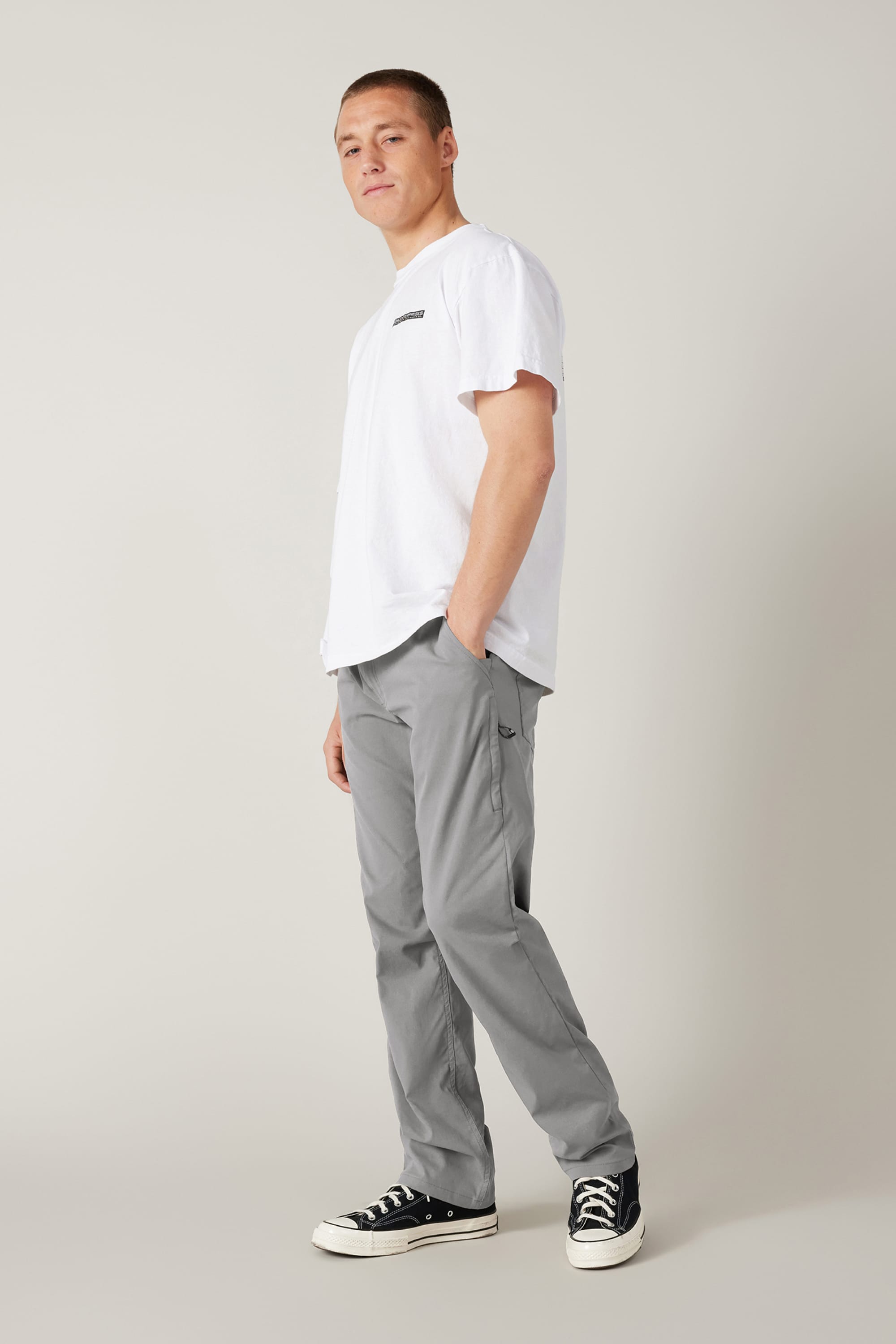 A person stands against a light gray background, wearing a white t-shirt, black-and-white sneakers, and 686 Men's Everywhere® Pant - Relaxed Fit from 686, with one hand in their pocket and a relaxed expression.