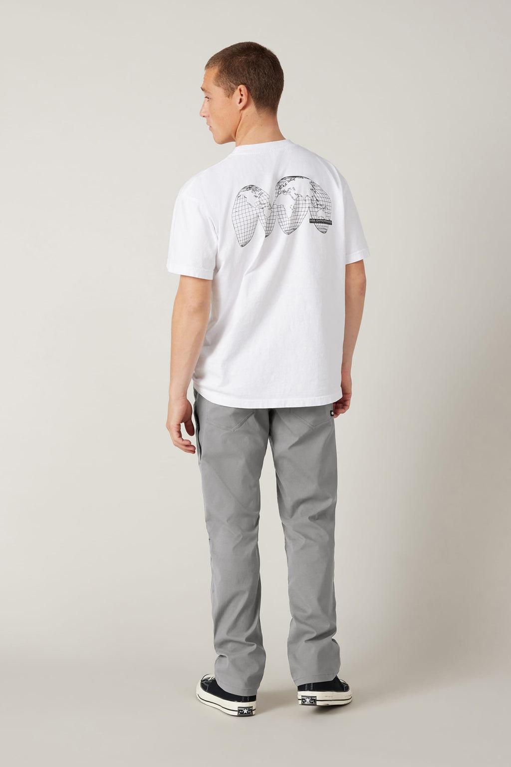 A man stands facing away, wearing a white T-shirt with three pineapples on the back, 686 Men's Everywhere® Pant - Relaxed Fit by 686 in breathable gray, and black-and-white sneakers, against a plain light background.