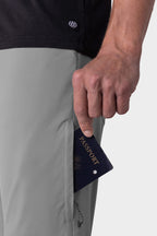 A hand slips a navy blue passport into the zippered pocket of light gray 686 Men's Everywhere® Pant - Relaxed Fit by 686, shown against a plain background.