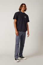 A young man with wavy hair wears a loose black t-shirt, 686 Men's Everywhere® Pant - Relaxed Fit by 686 in blue, and black-and-white sneakers while standing against a plain light background.