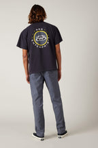 A person with shoulder-length brown hair faces away, wearing a black T-shirt with a circular graphic and text on the back, 686 Men's Everywhere® Pant - Relaxed Fit in blue by 686, and black-and-white sneakers against a plain background.