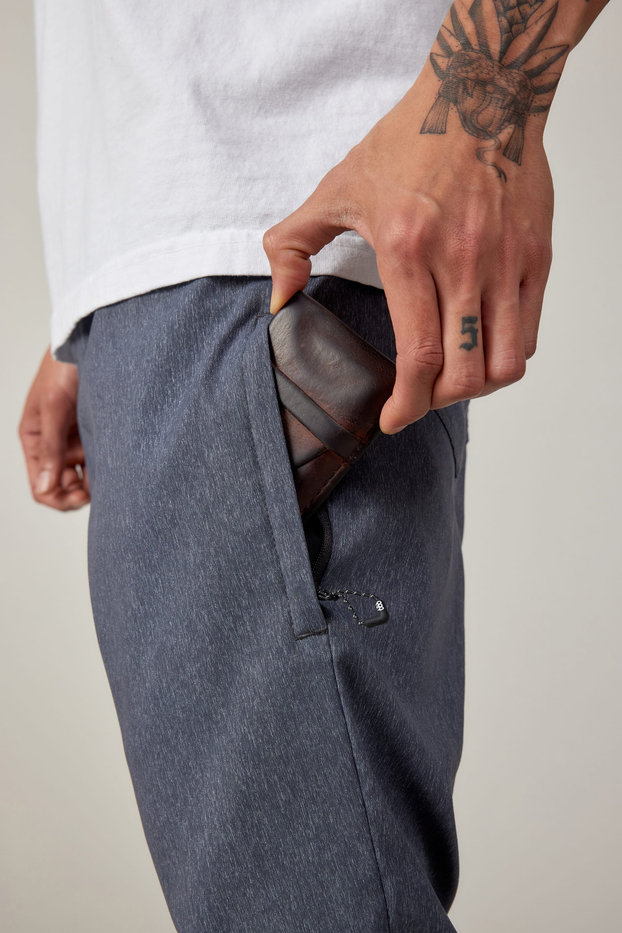 A person with tattoos on their hand puts a brown leather wallet into the zippered pocket of blue-gray 686 Men's Everywhere® Pant - Relaxed Fit, paired with a white shirt.