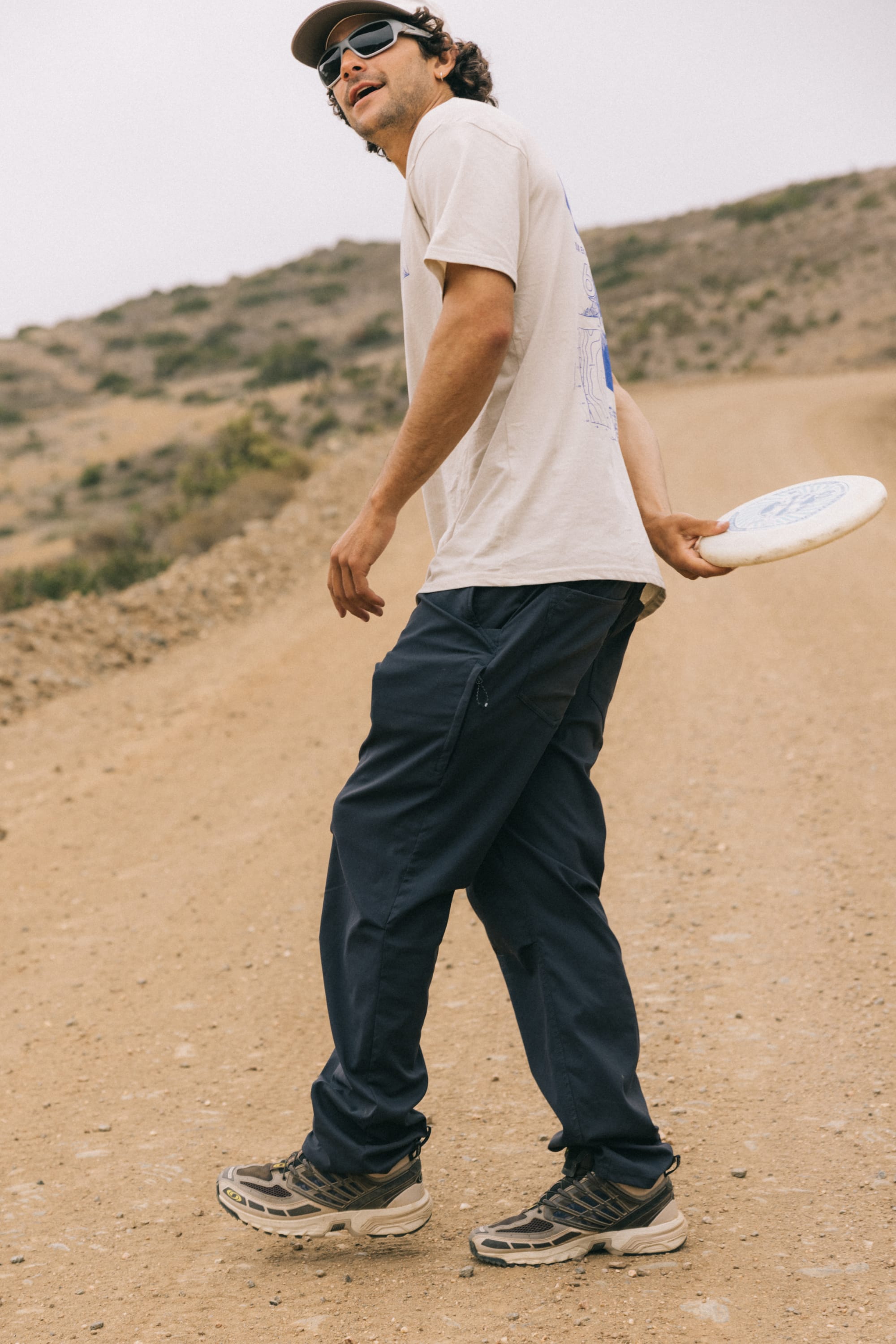 A person stands on a dirt road holding a frisbee behind their back, wearing sunglasses, a light t-shirt, sneakers, and 686 Men's Everywhere® Pant - Relaxed Fit by 686. The backdrop is hilly, dry terrain.