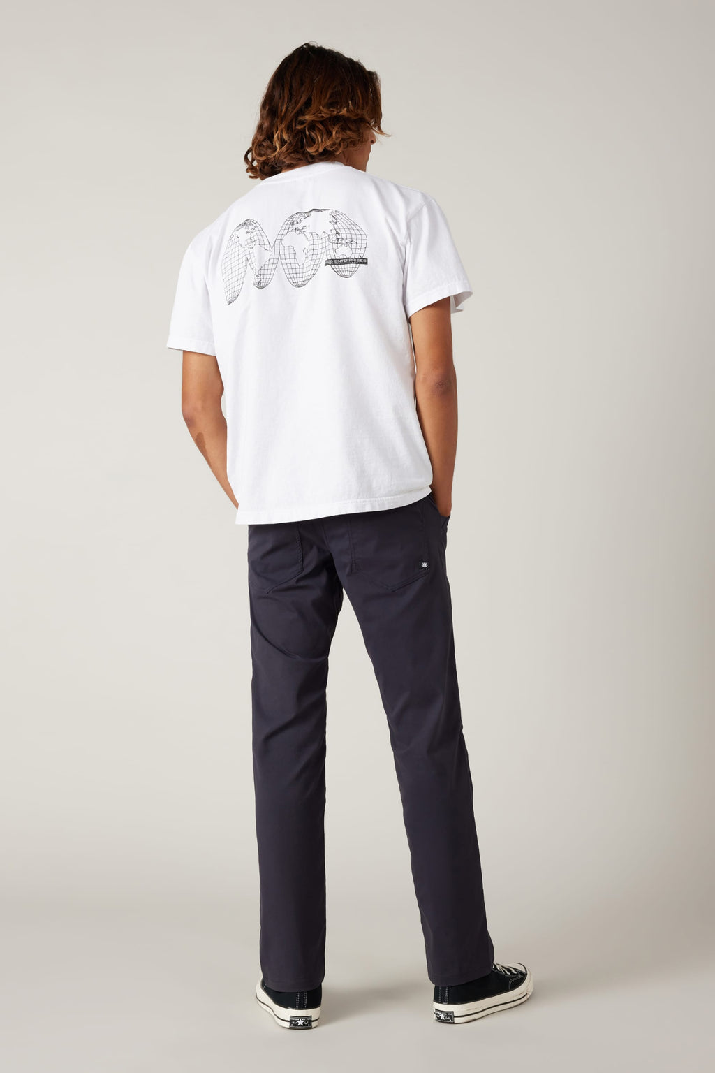 A person with shoulder-length hair stands facing away in a neutral studio, wearing a white tee with an abstract black back graphic, 686 Men's Everywhere® Pant - Relaxed Fit by 686, and black sneakers.