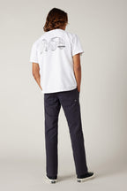 A person with shoulder-length hair stands facing away in a neutral studio, wearing a white tee with an abstract black back graphic, 686 Men's Everywhere® Pant - Relaxed Fit by 686, and black sneakers.