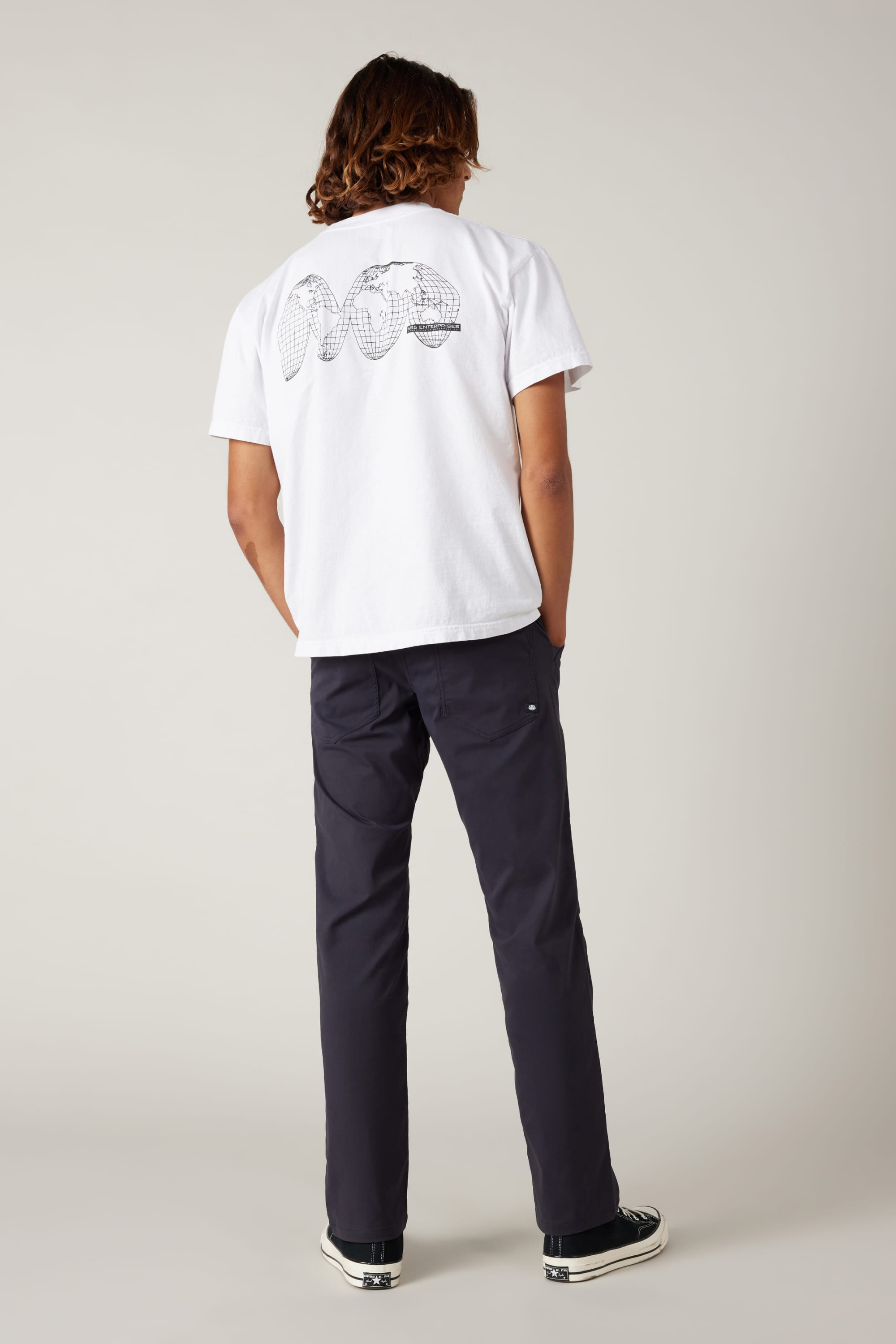 A person with shoulder-length hair stands facing away in a neutral studio, wearing a white tee with an abstract black back graphic, 686 Men's Everywhere® Pant - Relaxed Fit by 686, and black sneakers.