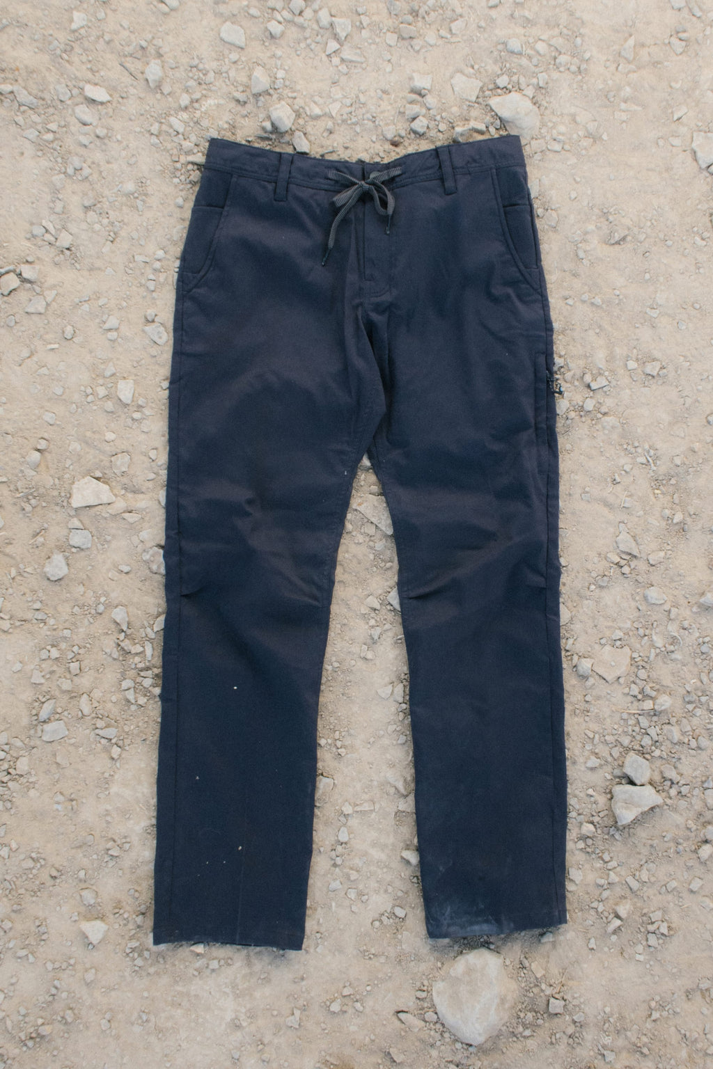 A pair of 686 Men's Everywhere® Pant - Relaxed Fit in dark blue, featuring a drawstring waist, lies flat on rocky, dirt-covered ground. The pants show slight wear at the cuffs.