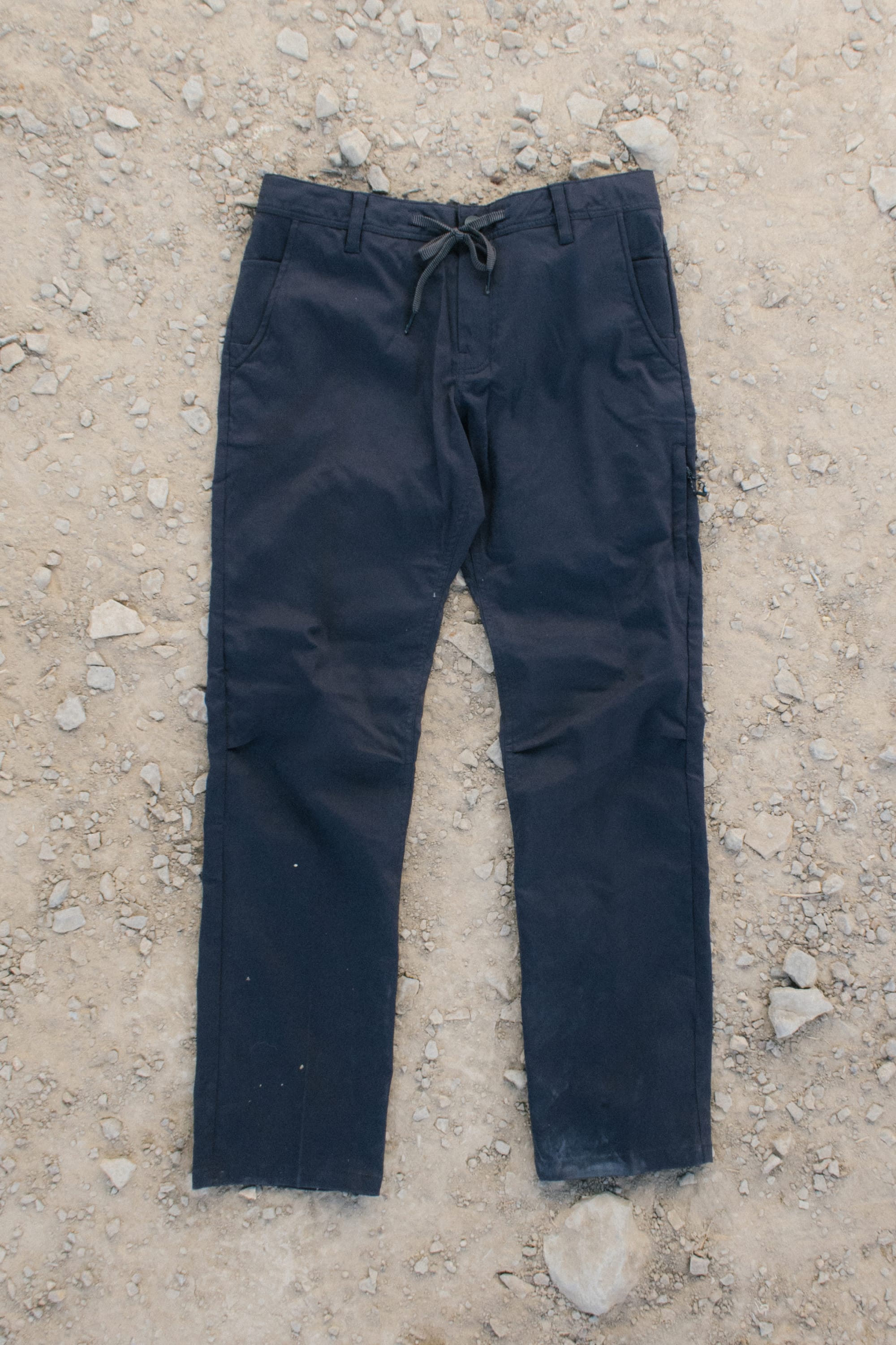 A pair of 686 Men's Everywhere® Pant - Relaxed Fit in dark blue, featuring a drawstring waist, lies flat on rocky, dirt-covered ground. The pants show slight wear at the cuffs.