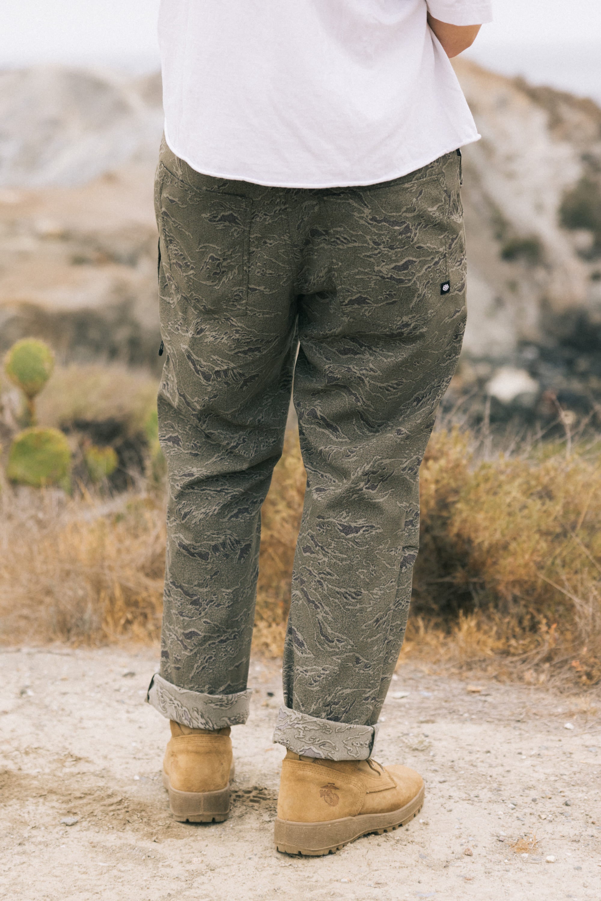 A person wearing 686 Men's Everywhere® Pant - Relaxed Fit in olive green, a white shirt, and tan suede boots stands outdoors on a dirt path with dry grass and a blurred rocky landscape, facing away from the camera.