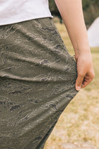 A person wearing 686 Men's Everywhere® Pant - Relaxed Fit in green camo gently pulls at the fabric outdoors on grass, with only their lower torso and hand visible.