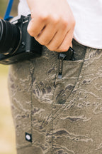 A person wearing 686 Men's Everywhere® Pant - Relaxed Fit holds a Gerber multitool clipped to their pocket, with a camera hanging from a strap around their neck.