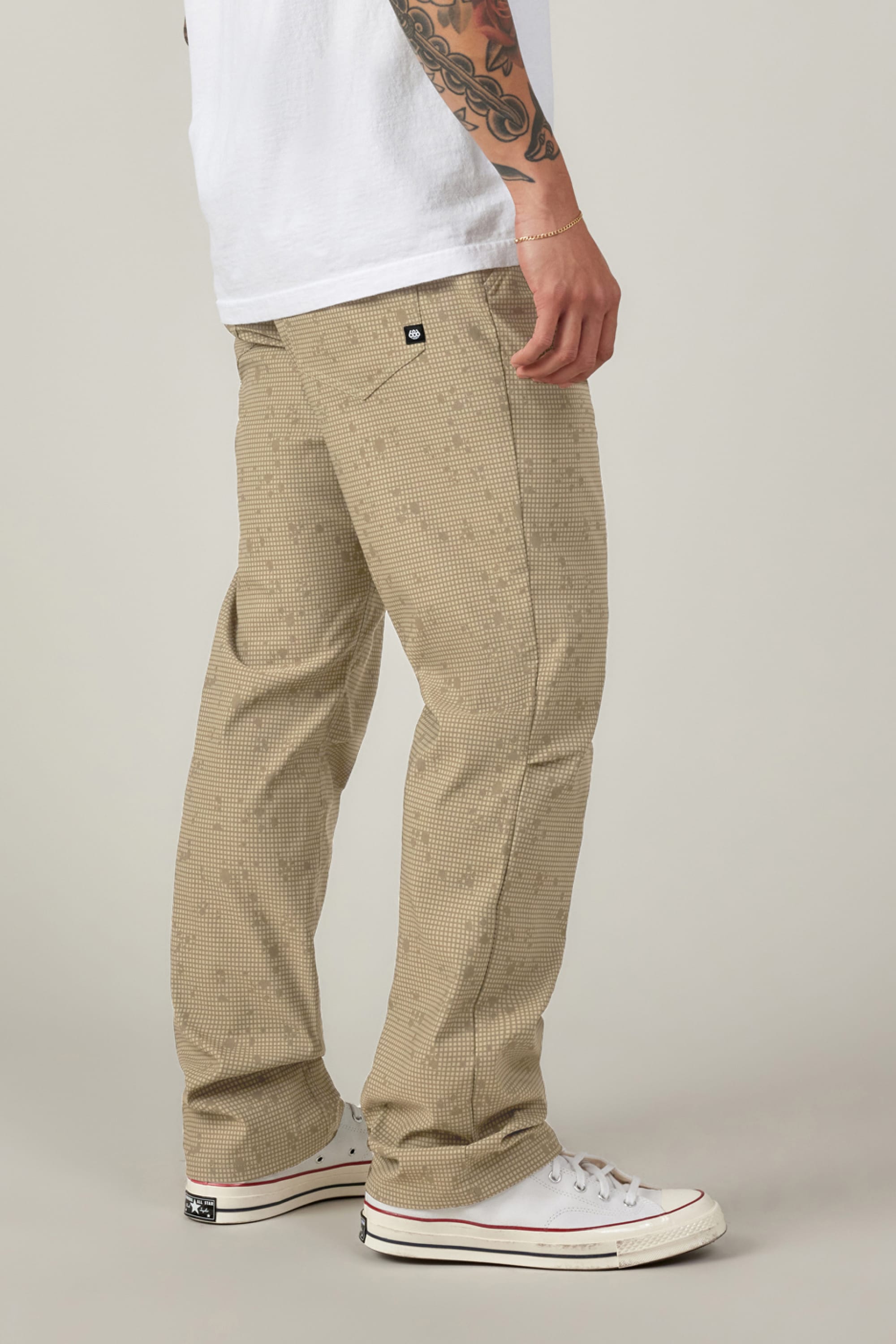 A person stands sideways against a plain light background, wearing 686 Men's Everywhere® Pant - Relaxed Fit in beige plaid, a white T-shirt, and white sneakers. Their left arm with a tattoo sleeve rests by their side.