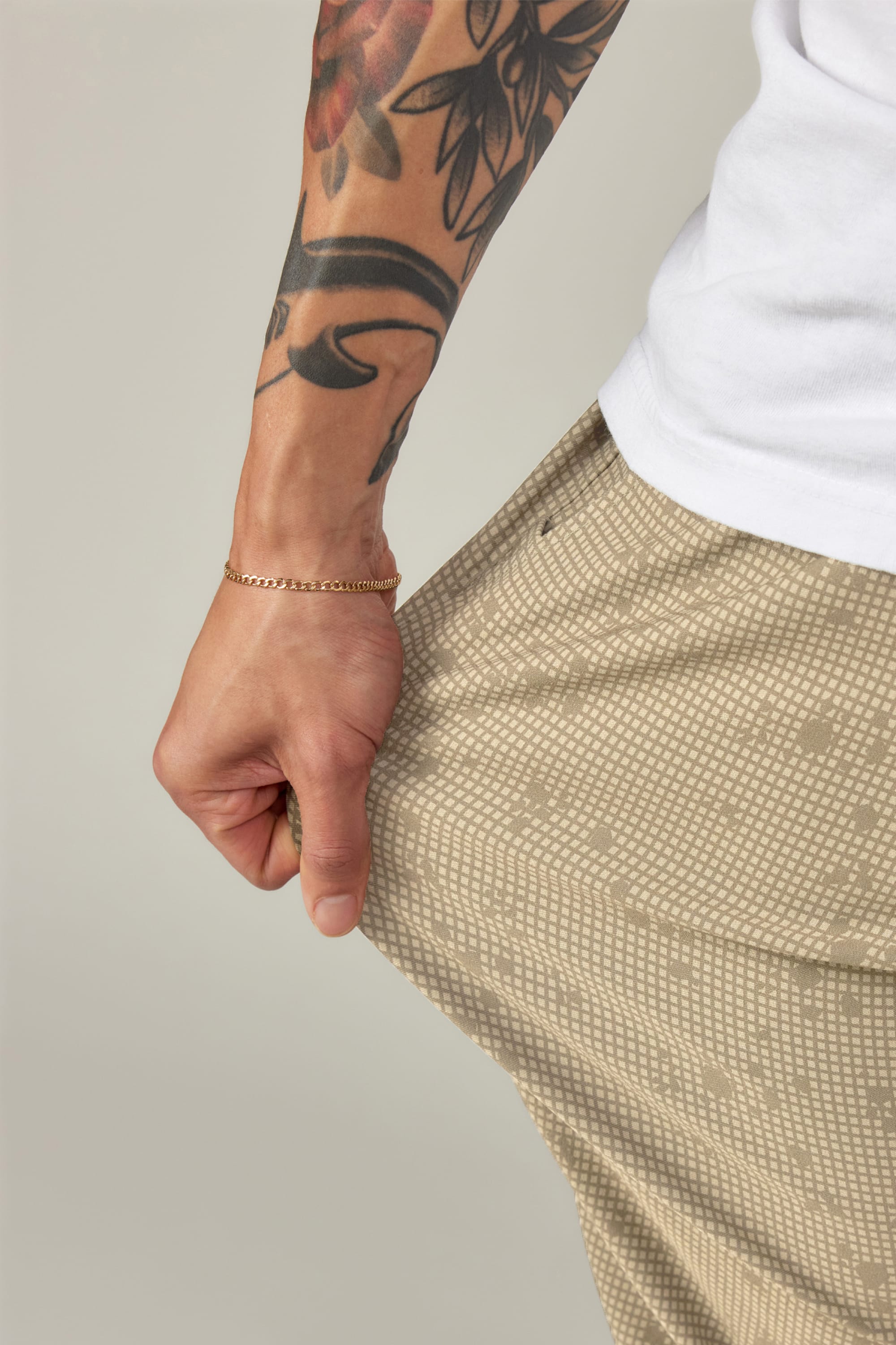 A person wears the 686 Men's Everywhere® Pant - Relaxed Fit in beige with a white shirt, showing a floral tattoo on their arm and a thin gold bracelet as they pull at their pocket.