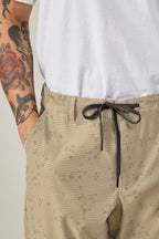 A person wears the 686 Men's Everywhere® Pant - Relaxed Fit in beige with a drawstring, paired with a white t-shirt. Their visible left arm has colorful tattoos, including a rose and a face.