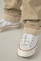 Close-up of a person wearing white lace-up sneakers and 686 Men's Everywhere® Pant - Relaxed Fit in beige with a grid pattern and red stitched rolled cuffs, blending comfort and style from 686.