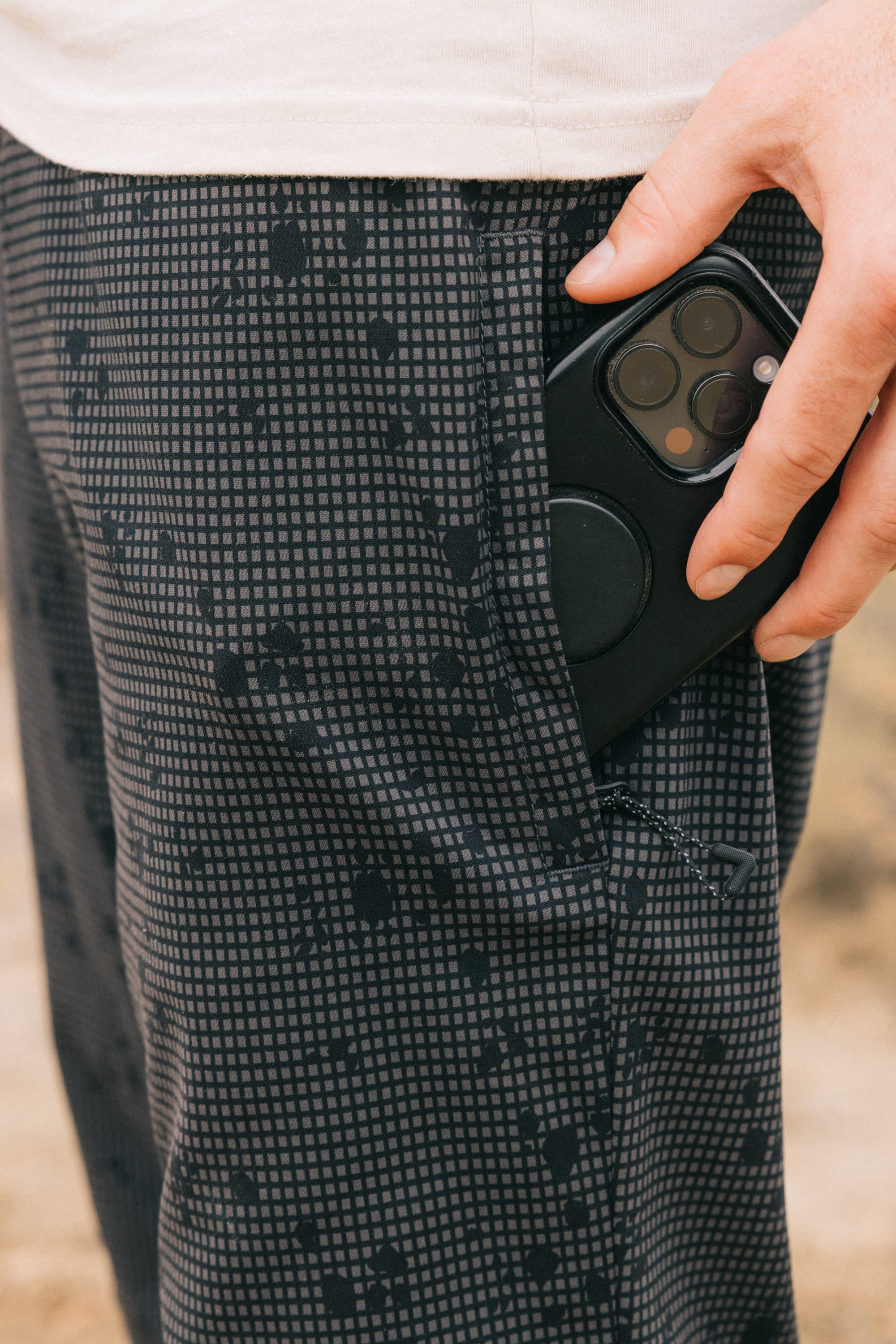 A hand places a black smartphone into the pocket of 686 Men's Everywhere® Pant - Relaxed Fit. The focus is on the hand, phone, and pocket, with a white shirt partially visible at the top.