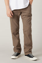 A person wearing 686 Men's Everywhere® Pant - Relaxed Fit by 686 in brown, a white T-shirt, and black low-top sneakers with white soles, stands against a plain light background.