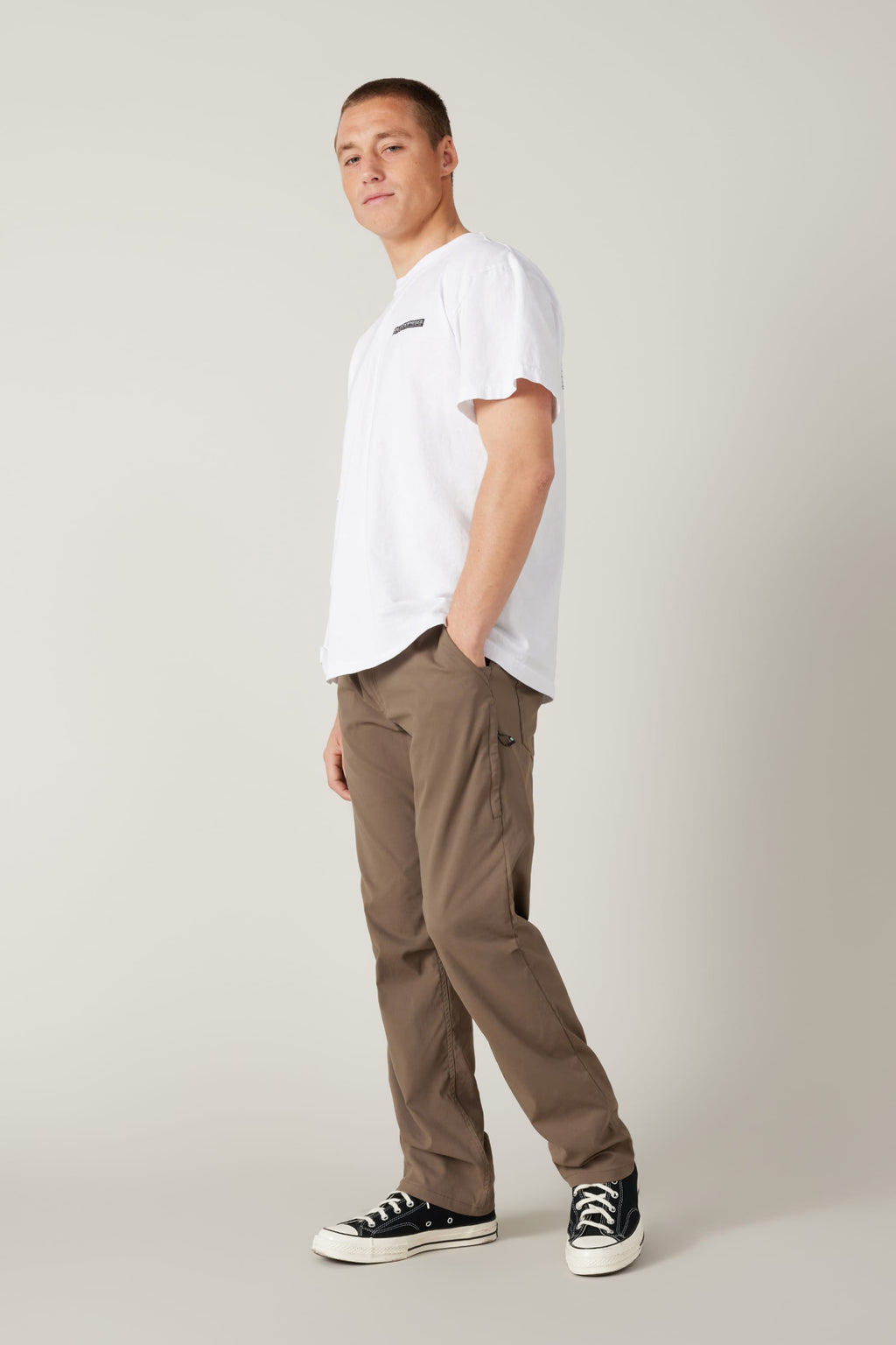 A person stands in a relaxed pose, wearing a white t-shirt, 686 Men's Everywhere® Pant - Relaxed Fit in brown, and black sneakers with white soles, against a plain light gray background.