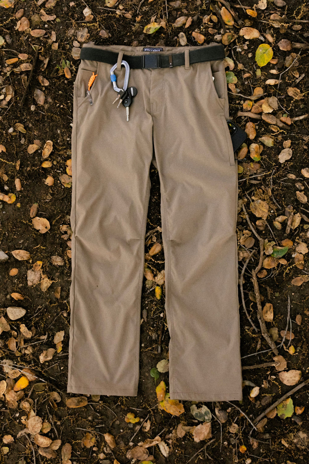 The 686 Men's Everywhere® Pant - Relaxed Fit in tan is shown laid flat on soil with fallen leaves and twigs, featuring a black belt, carabiner, keys, and pen attached to the belt loops.
