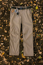 The 686 Men's Everywhere® Pant - Relaxed Fit in tan is shown laid flat on soil with fallen leaves and twigs, featuring a black belt, carabiner, keys, and pen attached to the belt loops.