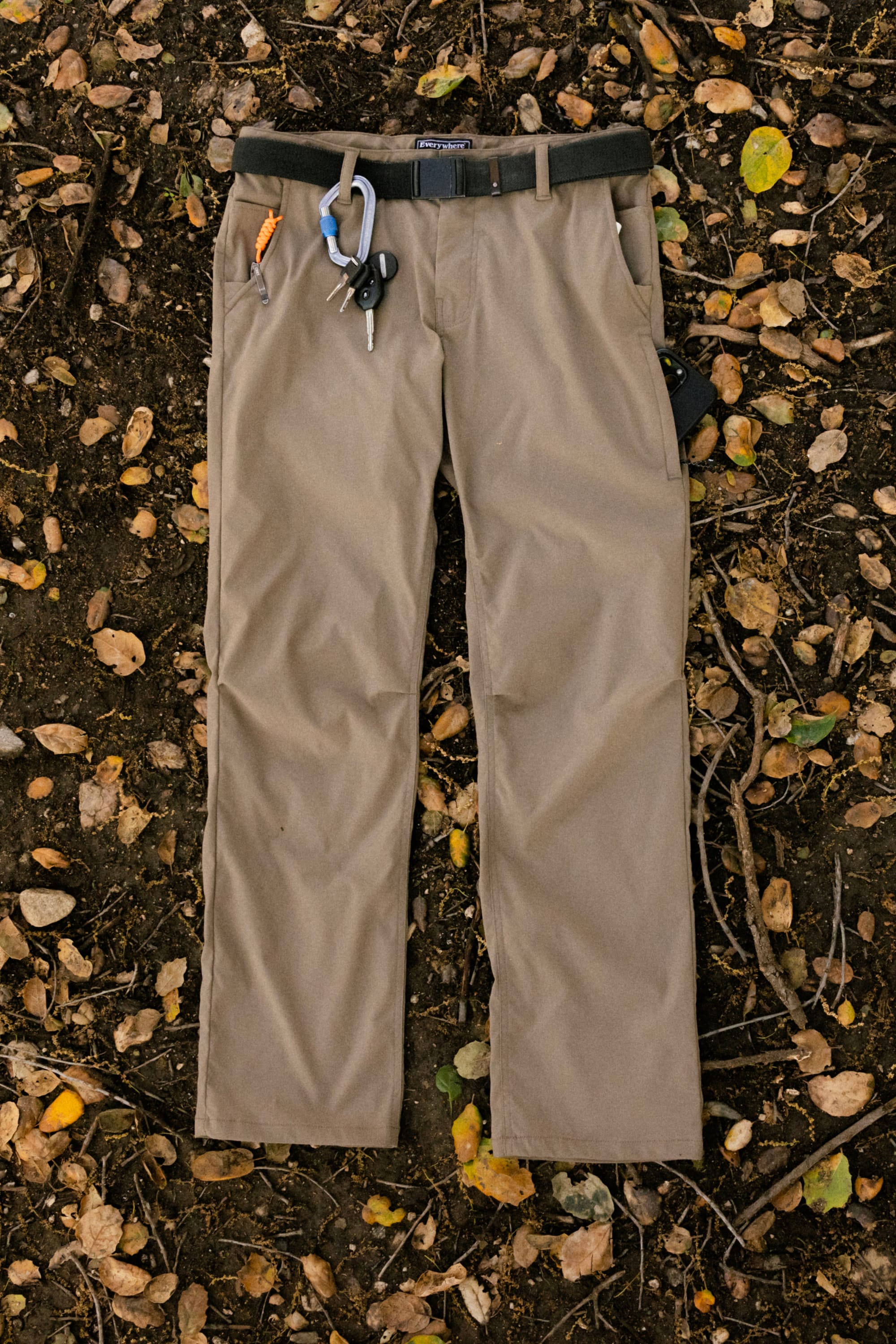 The 686 Men's Everywhere® Pant - Relaxed Fit in tan is shown laid flat on soil with fallen leaves and twigs, featuring a black belt, carabiner, keys, and pen attached to the belt loops.