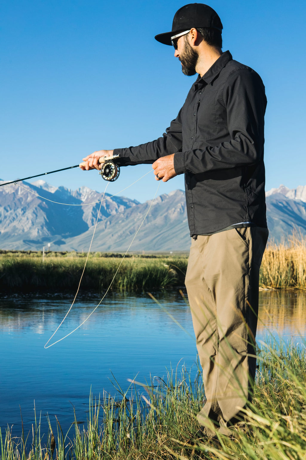 A man fly fishing in a river, wearing a black cap and shirt with 686 Men's Everywhere® Pant - Relaxed Fit, stands on a grassy riverbank with mountains and blue sky in the background.