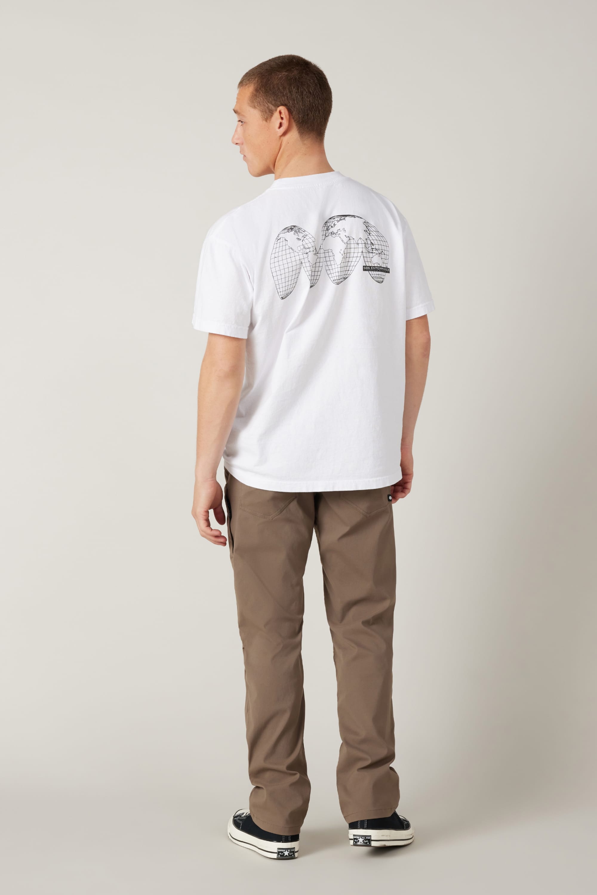 A man stands facing away, wearing a white tee with a graphic of three tennis rackets on the back, 686 Men's Everywhere® Pant - Relaxed Fit in brown, and black sneakers against a plain light background.