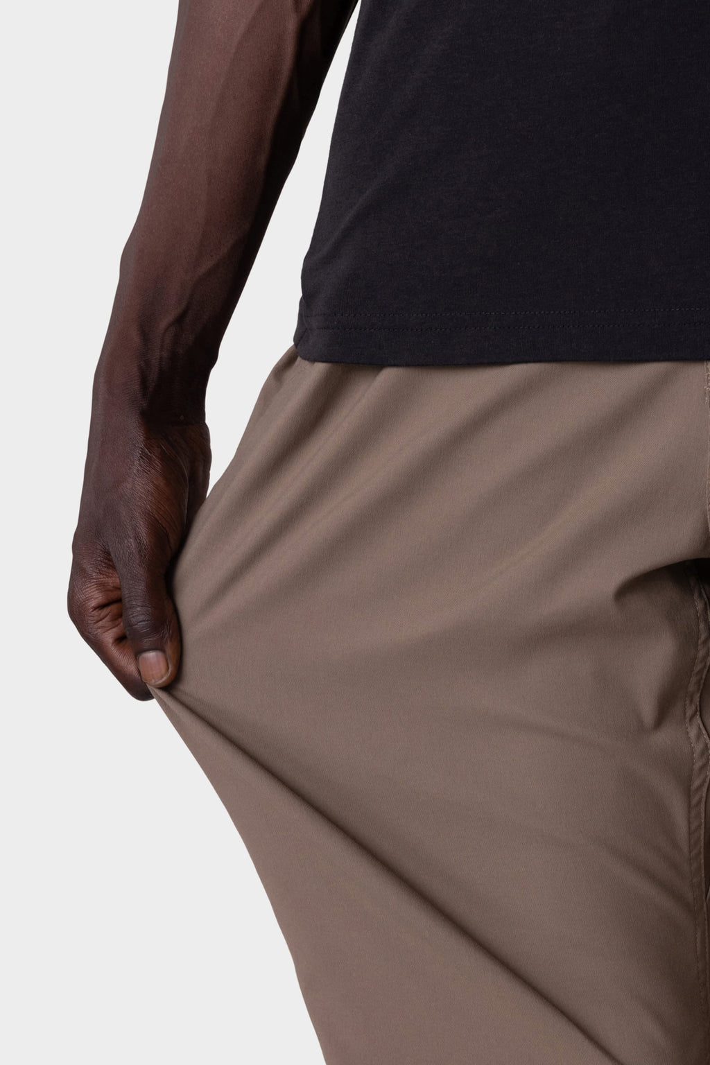 A person wears the 686 Men's Everywhere® Pant - Relaxed Fit in beige, stretching the fabric to show its flexibility while paired with a black shirt.