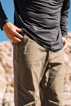 A person models the 686 Men's Everywhere® Pant - Relaxed Fit outdoors, their hand in a pocket. The blue sky and rocky terrain in the blurred background highlight the comfort and style of these 686 pants.