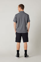 A man faces away in a short-sleeve gray button-up shirt and 686 Everywhere® Hybrid Shorts by 686, paired with black socks and black sneakers with white soles, against a plain light background.