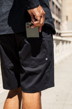 A person wearing 686 Everywhere® Hybrid Short and a dark jacket slips a smartphone into their back pocket outdoors on a sunny day, with only their lower body and hand visible.