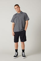 A man stands against a light gray background wearing a short-sleeve gray button-up shirt and 686 Everywhere® Hybrid Short in black, paired with black crew socks and sneakers. His left hand is in his pocket, with a relaxed expression.