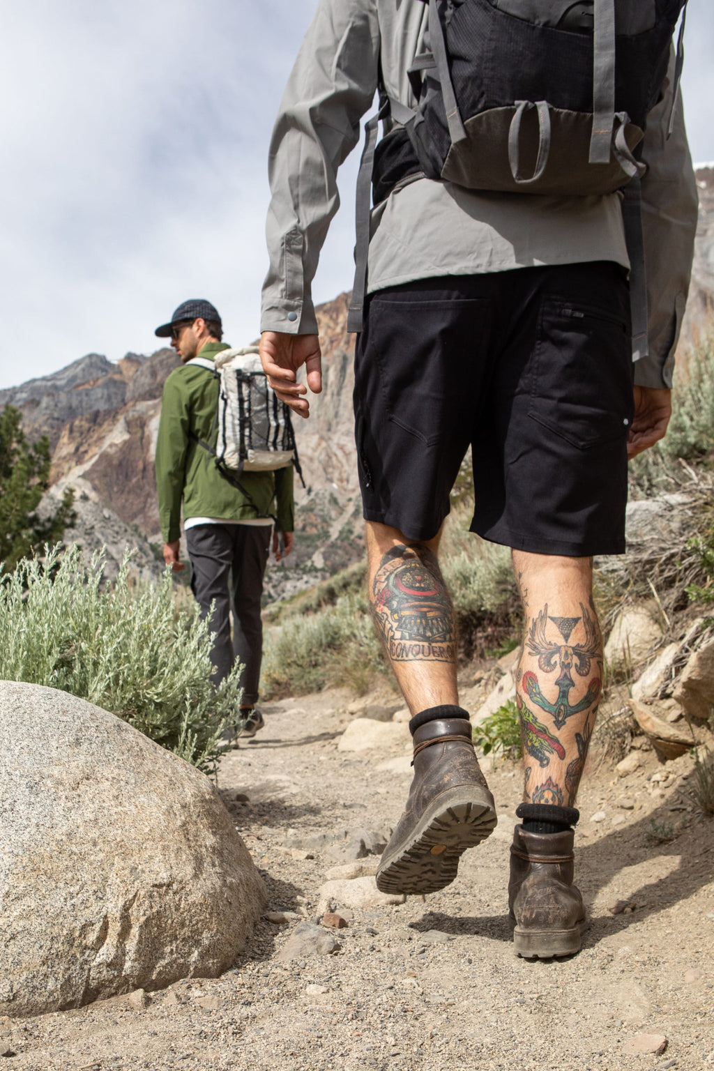 Two hikers trek a rocky trail with mountains and bushes. The nearer hiker wears 686 Everywhere® Hybrid Shorts by 686, showing leg tattoos, boots, and a backpack; the other, further ahead, sports long pants and a cap.