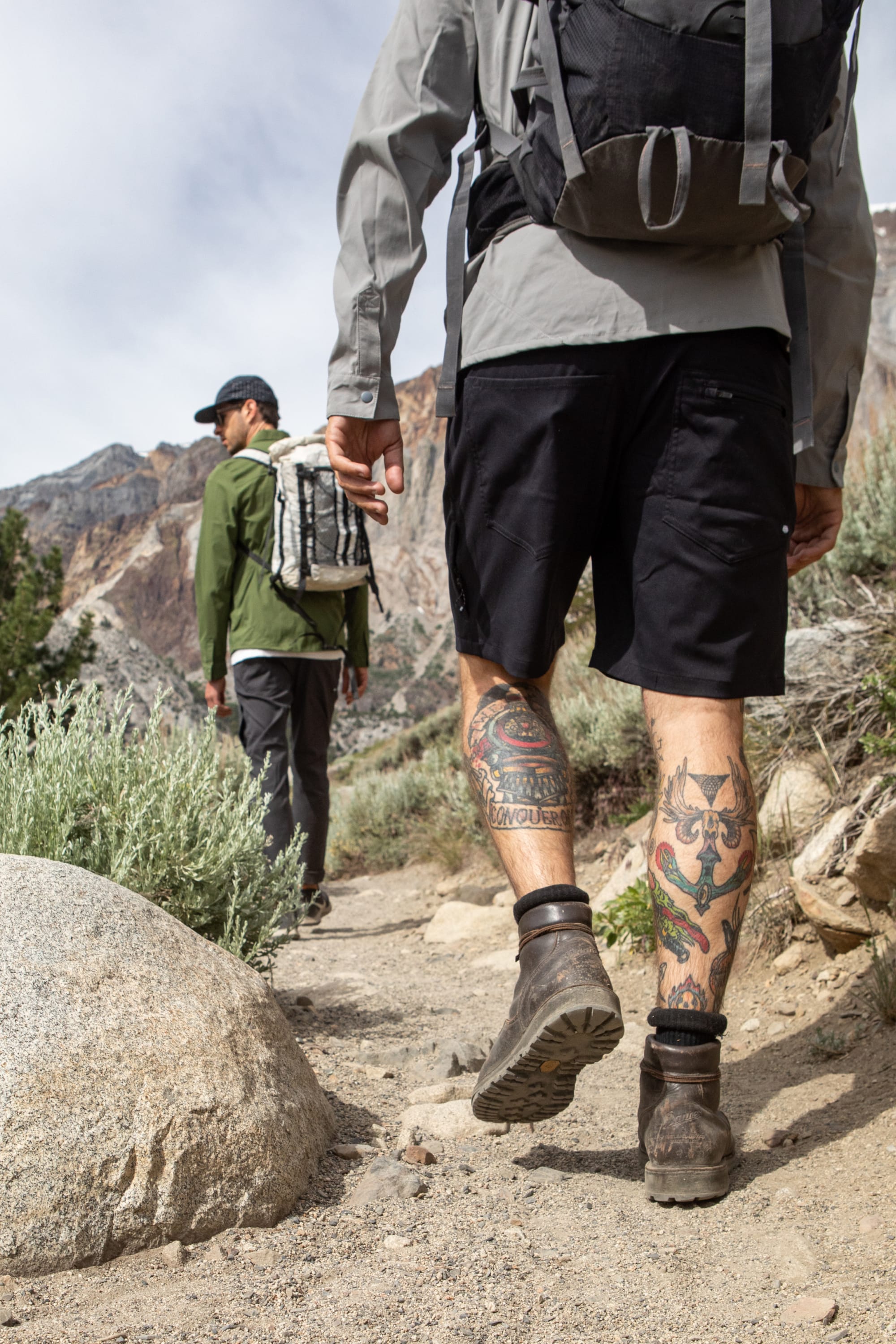 Two hikers trek a rocky trail with mountains and bushes. The nearer hiker wears 686 Everywhere® Hybrid Shorts by 686, showing leg tattoos, boots, and a backpack; the other, further ahead, sports long pants and a cap.