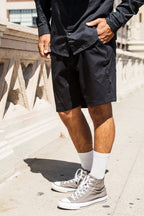A person stands outdoors on a sunny day wearing a black button-up shirt, 686 Everywhere® Hybrid Short by 686, white socks, and gray high-top sneakers, with one hand in their pocket next to a stone railing.