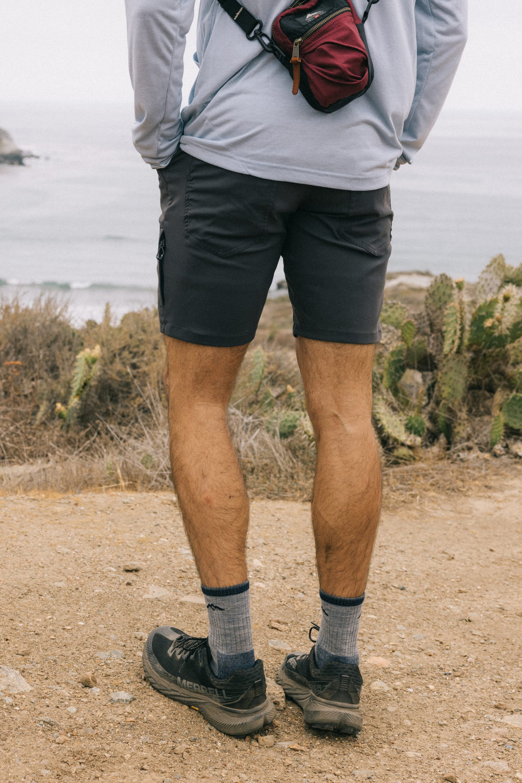 Wearing a breathable light long-sleeve shirt, 686 Men's Everywhere® Hybrid Short, hiking shoes, and a small red bag, a person stands on a dirt path by coastal vegetation, facing the ocean and distant cliffs.