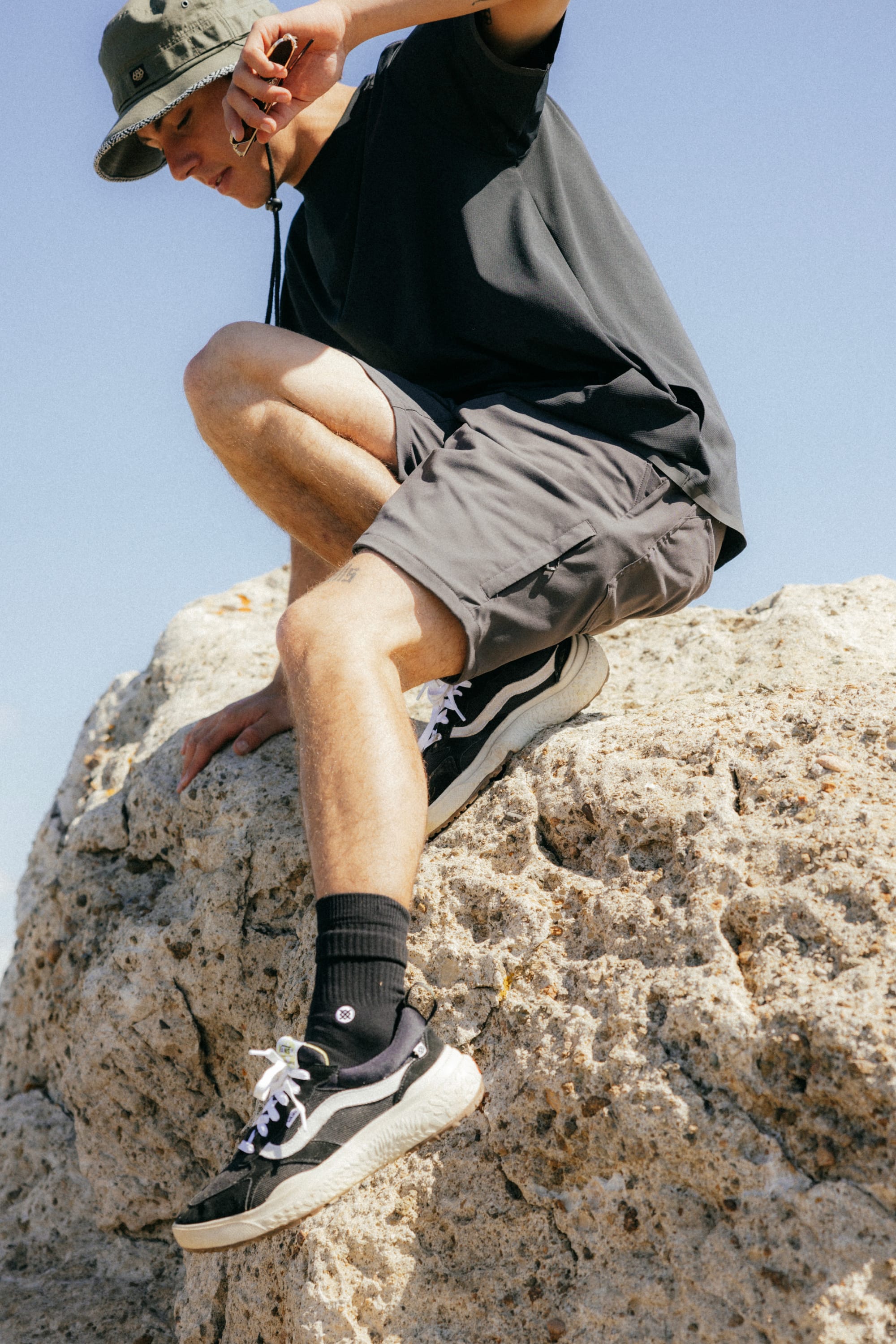 Wearing a bucket hat, dark t-shirt, 686 Everywhere® Hybrid Short, black socks, and black sneakers, a person climbs a large textured rock beneath a clear blue sky.