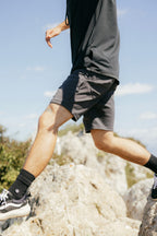 A person wearing 686 Everywhere® Hybrid Short, black shirt, black socks, and sneakers steps across rocky terrain on a sunny day. Only their lower body is visible.