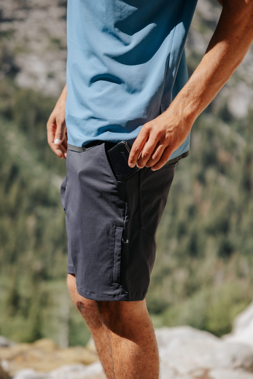 A person outdoors in a blue shirt and 686 Everywhere® Hybrid Short places a black smartphone into the zippered pocket, with blurred greenery and rocks in the background.