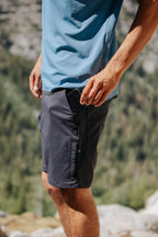 A person outdoors in a blue shirt and 686 Everywhere® Hybrid Short places a black smartphone into the zippered pocket, with blurred greenery and rocks in the background.