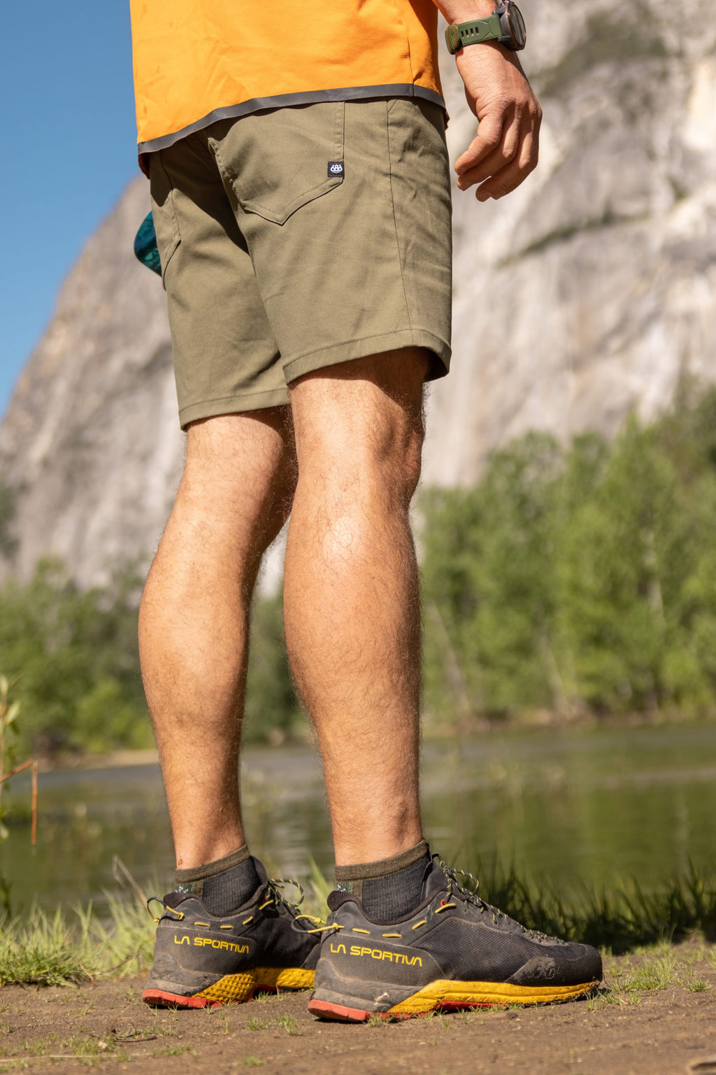 A person in an orange shirt, 686 Everywhere® Hybrid Short, and black La Sportiva hiking shoes stands outdoors by a river, with trees and a rocky cliff in the background.