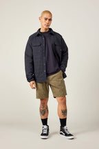 A person with short blond hair wears a black quilted jacket, dark shirt, black socks, black sneakers, and 686 Everywhere® Hybrid Short by 686 in olive green, standing against a plain background and showing leg tattoos.