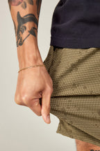 A close-up shows a tattooed forearm with a gold chain bracelet pulling the hem of 686 Everywhere® Hybrid Short, highlighting the green patterned, stain-resistant design and stylish practicality from 686.