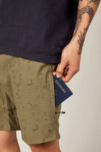 A person in a black shirt and patterned green 686 Everywhere® Hybrid Short holds a blue passport partly tucked in their pocket, with a visible tattoo on their arm.