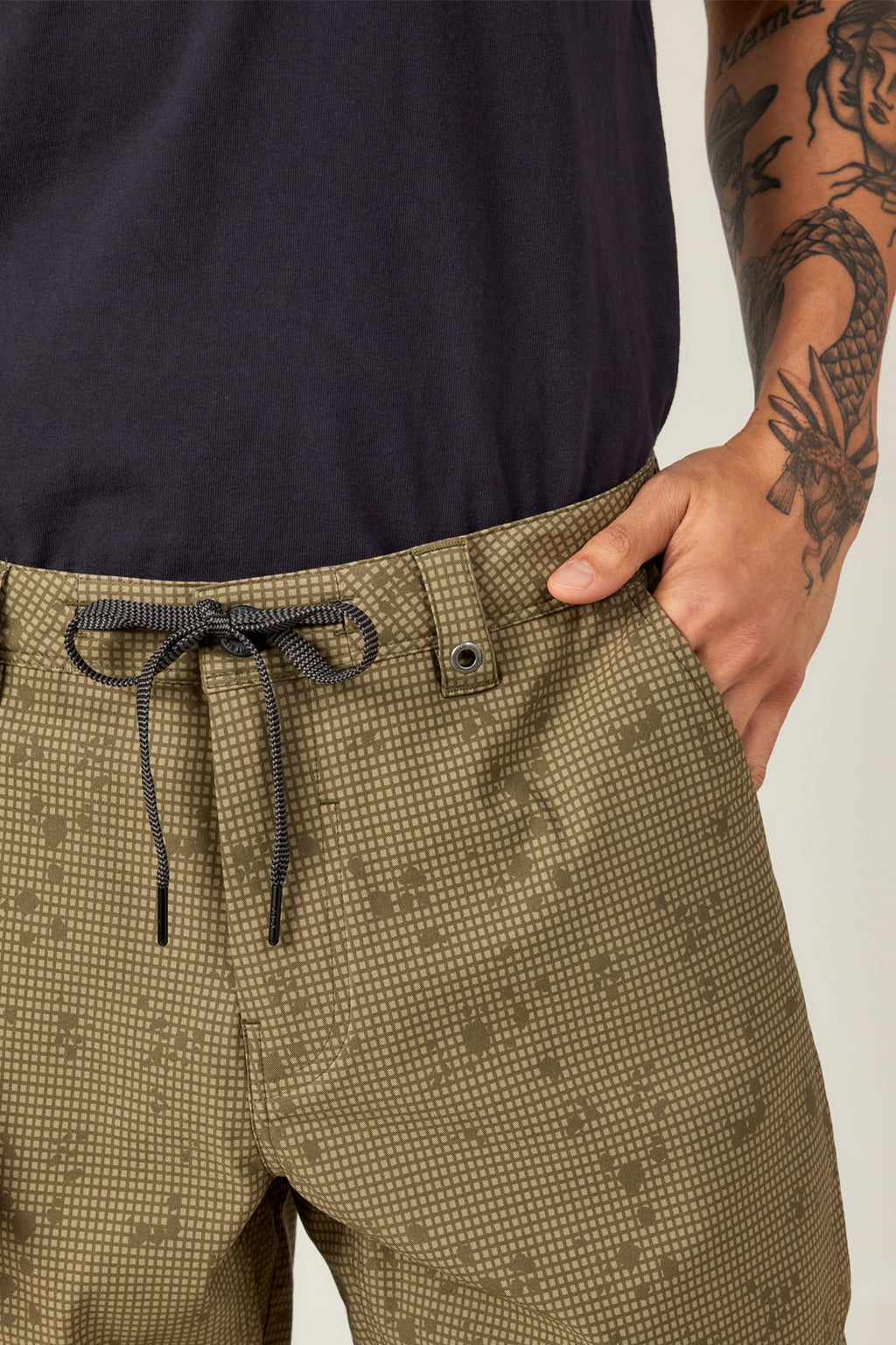Wearing light olive-green, patterned 686 Everywhere® Hybrid Shorts with a drawstring and black button, paired with a black shirt, a person rests their right hand in the pocket, showing a dragon tattoo on their forearm.